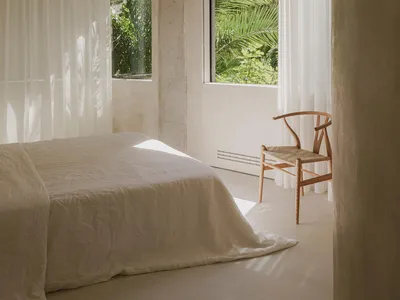 A minimalist bedroom with natural fibers, featuring a bed with white sheets, a wooden chair, and sheer curtains.