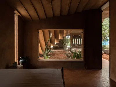 A modern bedroom featuring textured clay walls, concrete floors, and a window overlooking a courtyard with plants.