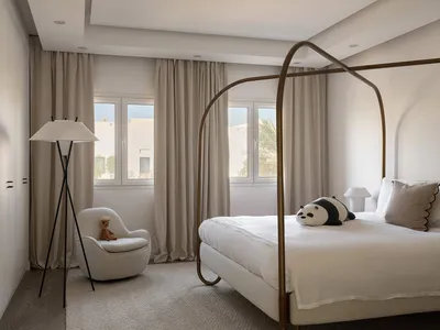 Modern bedroom featuring a fabric and metal bed frame, large windows with beige curtains, and a white upholstered chair with
