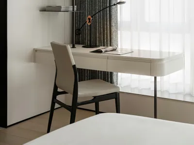 A minimalist bedroom workspace with a white desk and chair, a modern lamp, and a window with gray curtains.
