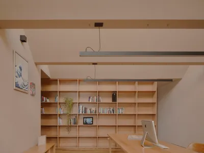 A minimalist home office with wooden shelves filled with books and plants, featuring a desk with a computer and keyboard.