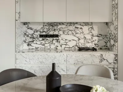 A modern kitchen featuring marble countertops and cabinets against a white marble backsplash, with a sleek vase and bowl of f