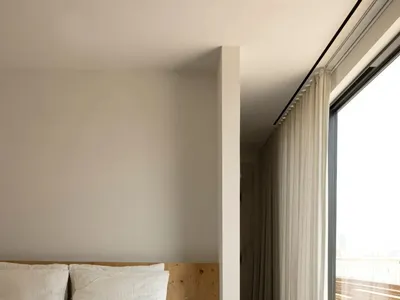 A minimalist bedroom with a wooden bed platform, concrete floor, and large window with sheer curtains.
