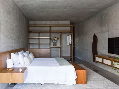 A minimalist bedroom with a concrete wall and wooden furniture, featuring a bed with white linens and a built-in wardrobe.