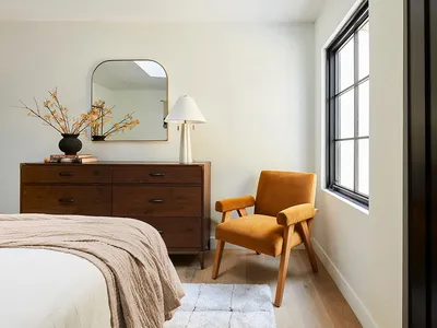A minimalist bedroom with a wooden dresser, orange chair, and white bedding.