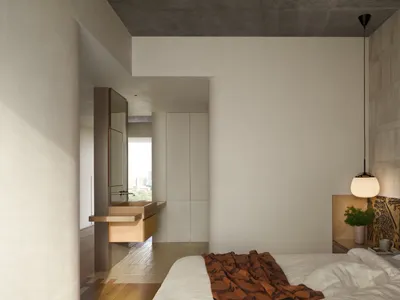 A minimalist bedroom with concrete and wooden elements, featuring a bed with a blanket and a hanging lamp.