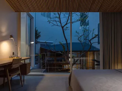 A minimalist bedroom with a wooden bed, desk, and ceiling design, featuring a large window overlooking a courtyard with trees