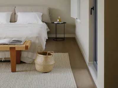 A minimalist bedroom with a neutral color palette, featuring a bed, side table with a bottle, and woven basket on a light-col