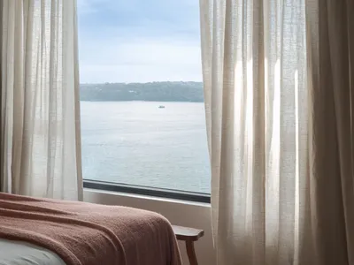 A bedroom with a bed covered in a reddish-brown blanket, floor-to-ceiling curtains with sheer white fabric, and a view of a c