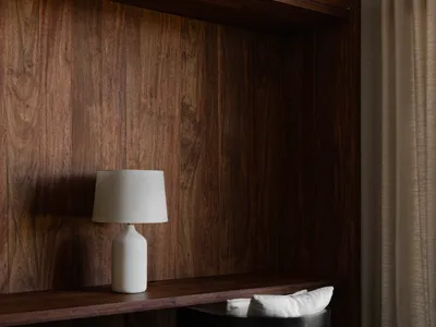 A simple home office featuring a dark wood desk, a black chair with a white cushion, a white table lamp, and wood-paneled wal