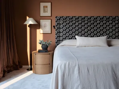 A modern bedroom with a patterned upholstered headboard, white bedding, a wooden bedside table, a floor lamp, and a framed ar