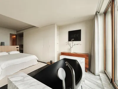 A minimalist bedroom featuring a large bed with a wooden bed frame, marble flooring, and a glass-enclosed bathtub.