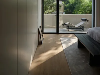 A minimalist bedroom with a bed, black bench, and a view of a balcony with a concrete wall and greenery.