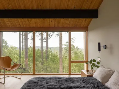 A minimalist bedroom with a large window overlooking a forest, featuring a bed with a grey blanket and a light brown leather