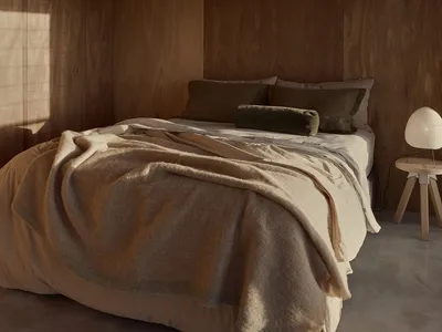 A minimalist bedroom featuring wooden walls, a bed with cream-colored bedding, and a small wooden side table with a lamp.