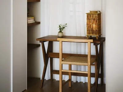 A minimalist home office with a wooden desk, chair, and a lantern lamp, featuring sheer curtains and a bookshelf.