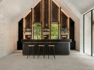 A modern bar interior featuring a concrete and brass counter with three stools, set against a textured wall and glass wall.