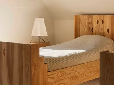 A minimalist bedroom featuring a wooden bed frame with geometric cutouts, a matching wooden nightstand, and a white lamp with