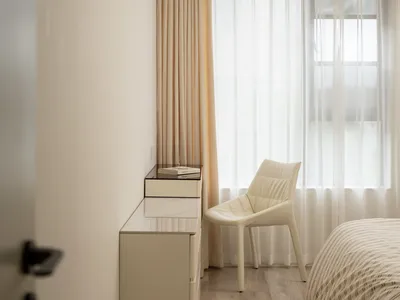 A minimalist bedroom with white furniture, featuring a bed with striped bedding and light-colored curtains.