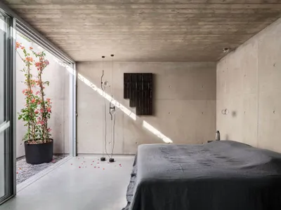 A minimalist bedroom featuring a concrete floor and walls, with a grey bed and geometric lighting fixtures, and a potted plan
