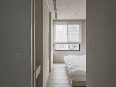 A minimalist bedroom featuring light-colored wood flooring, white bedding, and a large window with blinds.