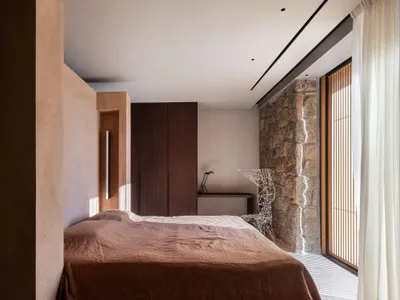 A minimalist bedroom featuring wooden furniture and a stone wall, with natural light streaming through the window.