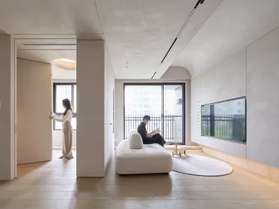 A minimalist living room with concrete walls and a white sofa, featuring a woman opening a door leading to a bedroom and a ma