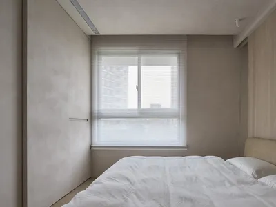 A minimalist bedroom with neutral-toned walls, a bed with white bedding, and white blinds on the window.
