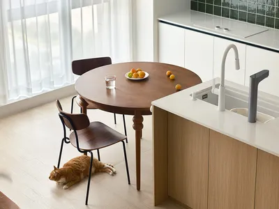 A minimalist residential kitchen featuring a round wooden table with fruits, a white countertop, and white cabinetry.