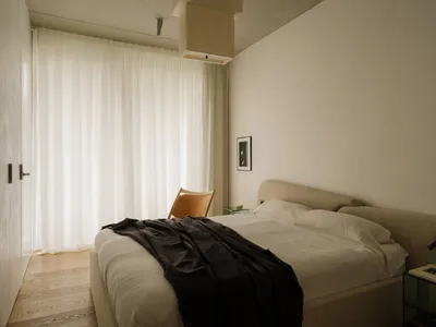 Modern minimalist bedroom with fabric bed, leather chair, and white curtains.