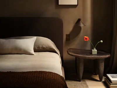 A minimalist bedroom featuring a bed with a beige comforter and white pillows, a wooden bedside table with a vase of flowers,