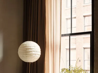 A modern home office featuring a wooden desk with a white paper lantern lamp, brown curtains, and a vase with flowers.