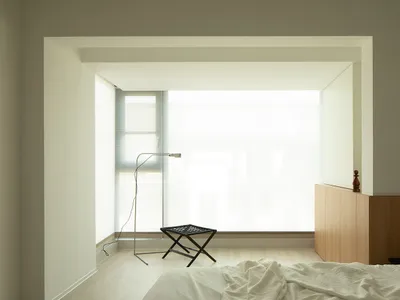 A modern minimalist bedroom with a large window, natural light, a bed, and a black ottoman.
