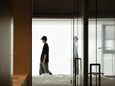 A minimalist bedroom with a bed covered in white linen, natural light streaming in through the window, and a person walking i