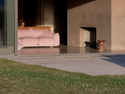A minimalist living room with a concrete exterior wall, featuring a pink sofa, a fireplace, and a wooden stool.