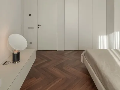 Modern minimalist bedroom with white walls, wooden flooring, and a single white lamp on a floating nightstand.