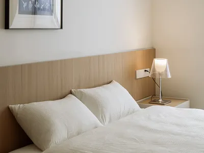 A minimalist bedroom with a white bed, lamp, and wooden headboard.