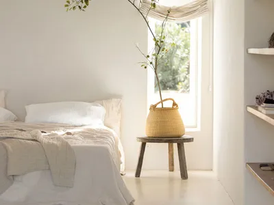 A minimalist bedroom with neutral-toned bedding, a wooden stool, and a woven basket with a plant.