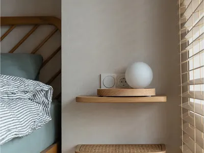 A minimalist bedroom featuring floating shelves made of wood, with a white spherical lamp, electrical outlets, and a bed with