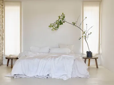 A minimalist bedroom featuring a bed with white cotton bedding, two windows with sheer curtains, and rustic wooden stools wit
