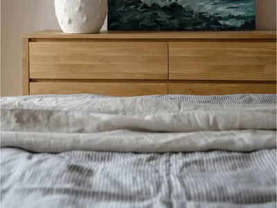 A bedroom with a wooden dresser, a green plant in a white vase, and a painting of a person in the ocean.