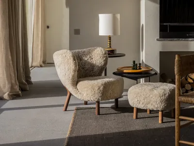 A cozy living room with a sheepskin chair and ottoman, a side table with a lamp, and a fireplace.