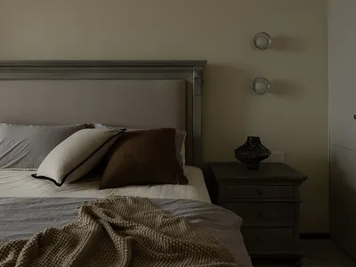 A minimalist bedroom featuring a bed with a knitted blanket, a headboard, and a bedside table with decorative items.