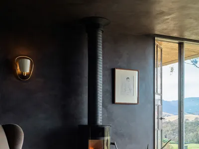 A cozy industrial-style living room features a concrete fireplace, wood floors, and a rustic rug, with a view overlooking a g