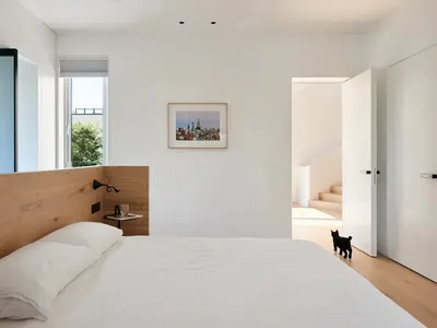 Modern minimalist bedroom with wooden headboard, white bedding, cityscape artwork, and black cat.