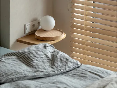 A minimalist bedroom with a wooden blind, grey sheets, and a round wooden tray with a white globe lamp.