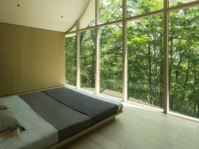 A modern minimalist bedroom with a large window showcasing a forest view, featuring wooden flooring and a low-profile bed.