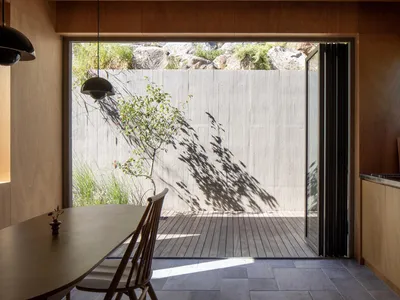 A minimalist residential kitchen featuring a wooden floor and a plant casting shadows on a wall.