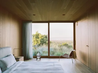A minimalist bedroom featuring a bed with a wooden headboard and floor, large windows overlooking the landscape, and a wooden