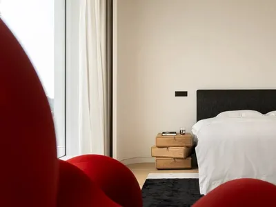 Modern minimalist bedroom featuring minimalist white fabric furnishings on a wooden bed platform, with a red curved chair and